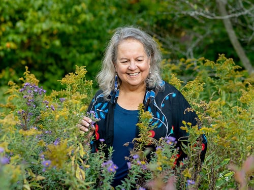 Robin Wall Kimmerer smiles among purple and yellow wildflowers, gently holding a flowering stem with leafy trees behind her.