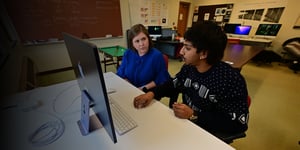 A student and professor work together at a desktop computer in a campus computer lab.