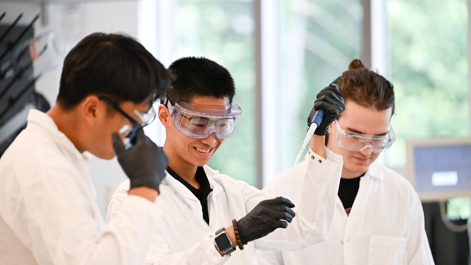 Three students in lab coats and safety goggles use pipettes in a bright lab, smiling and focused as they conduct a hands-on science experiment together.