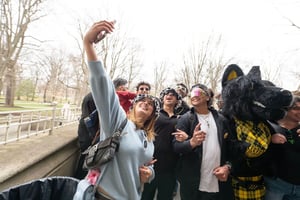 A group of students crowd together outdoors, smiling and posing for a selfie on a campus walkway. One person holds a phone up high while Archie, Wooster's mascot, in a yellow tartan outfit stands beside them.