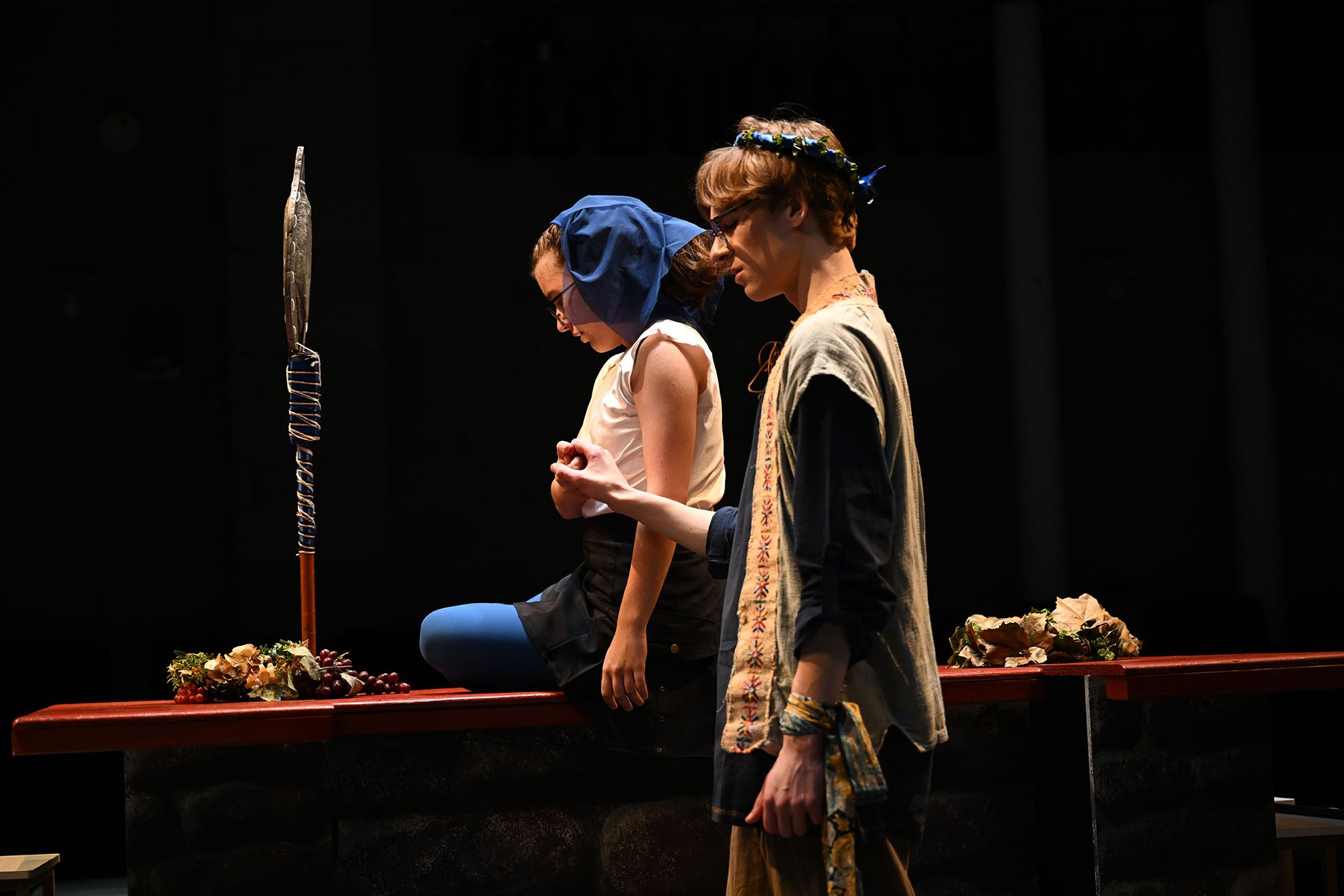 Two students in period costumes perform on a dimly lit theater stage beside a long table decorated with grapes and autumn leaves, with one kneeling in reflection and the other walking past wearing a floral wreath.