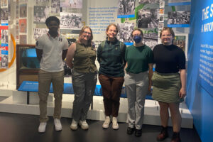 This image shows a group of five people standing side by side in front of an exhibit wall. The wall behind them displays various historical photos and text about social justice.