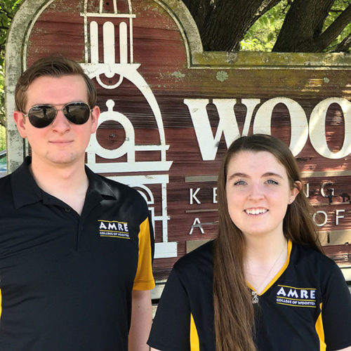two students wearing black polos pose in front of College of Wooster sign
