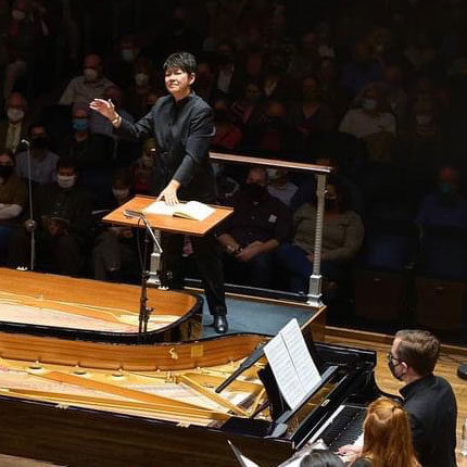 Lisa Wong associate professor of music at Wooster and director of choruses at The Cleveland Orchestra standing on a stage directing a chorus with two musicians seated at a piano in the lower right corner