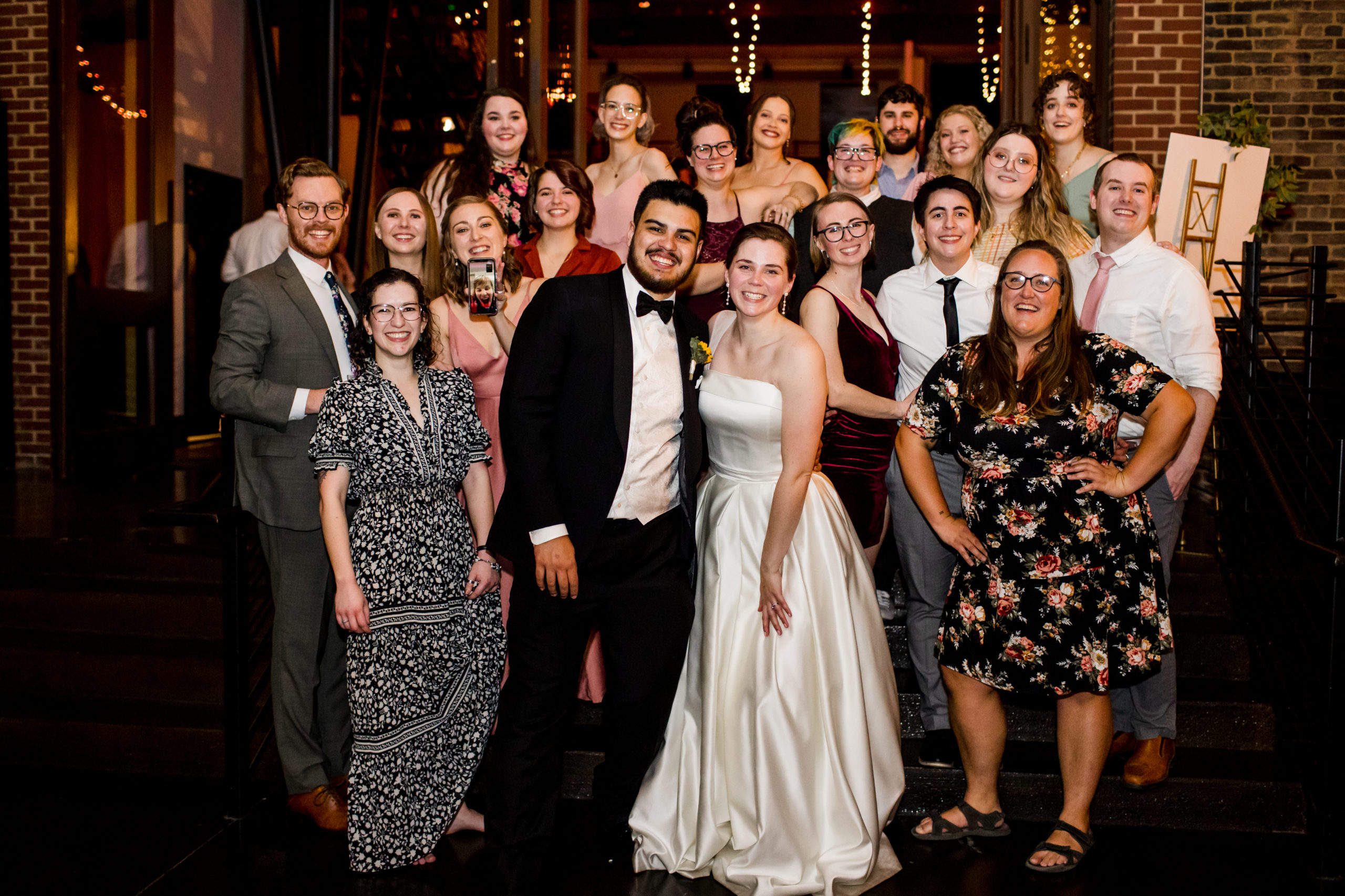 20 wedding members smile while posing for Charlotte Tierney and Connor Burnard's marriage. One member holds the phone on a video call for a virtual member to pose as well, making it 21 members. Front and Center are the bride and groom.