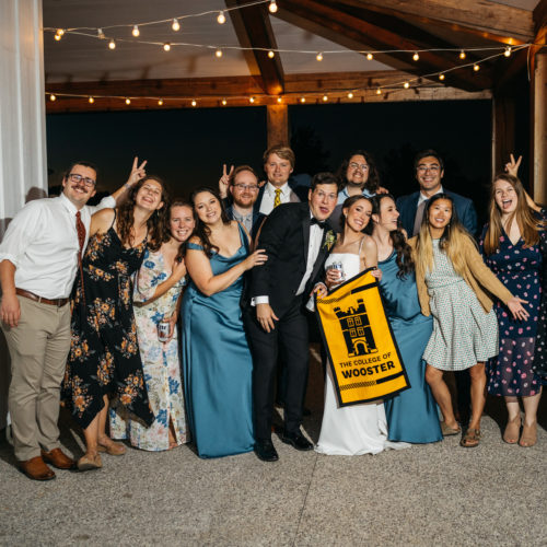 14 wedding members pose to commemorate Allie Elchert ('17) and Tanner Fisher's marriage. The bride and groom are in the center of the group holding a College of Wooster felt banner.