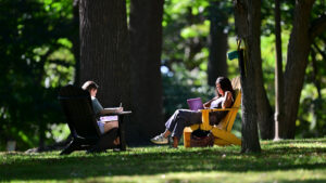 Two students at The College of Wooster sit outdoors in Adirondack chairs on a grassy area beneath large trees. One student, seated in a black chair, is writing or sketching in a notebook, while the other, in a yellow chair, is focused on a laptop. The setting is peaceful, with sunlight filtering through the trees, creating a serene and focused atmosphere for studying or working in nature.