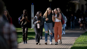 Group of Wooster students walking on a sunny path, looking at their phones and laughing, while others walk in the background.