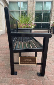 Black metal bench with plaque reading "In memory of Nan Nichols, Pioneer in Women's Athletics," placed on a brick walkway with planters and a building in the background.