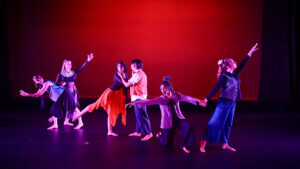 "Six dancers perform on stage under red lighting, striking expressive poses in a contemporary piece with flowing costumes and dramatic movement.