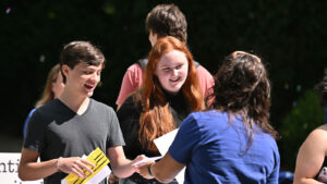 Smiling students at The College of Wooster interact outdoors during an event, holding papers and talking with a staff member in a blue shirt.
