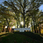 Sunlight filters through tall oak trees surrounding the College of Wooster entrance sign, which reads “College of Wooster — Founded 1866,” on a bright autumn day.