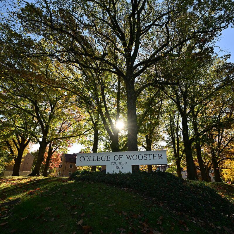 Sunlight filters through tall oak trees surrounding the College of Wooster entrance sign, which reads “College of Wooster — Founded 1866,” on a bright autumn day.